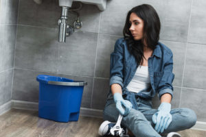 Woman sitting on bathroom floor beside leaking pipe with blue bucket